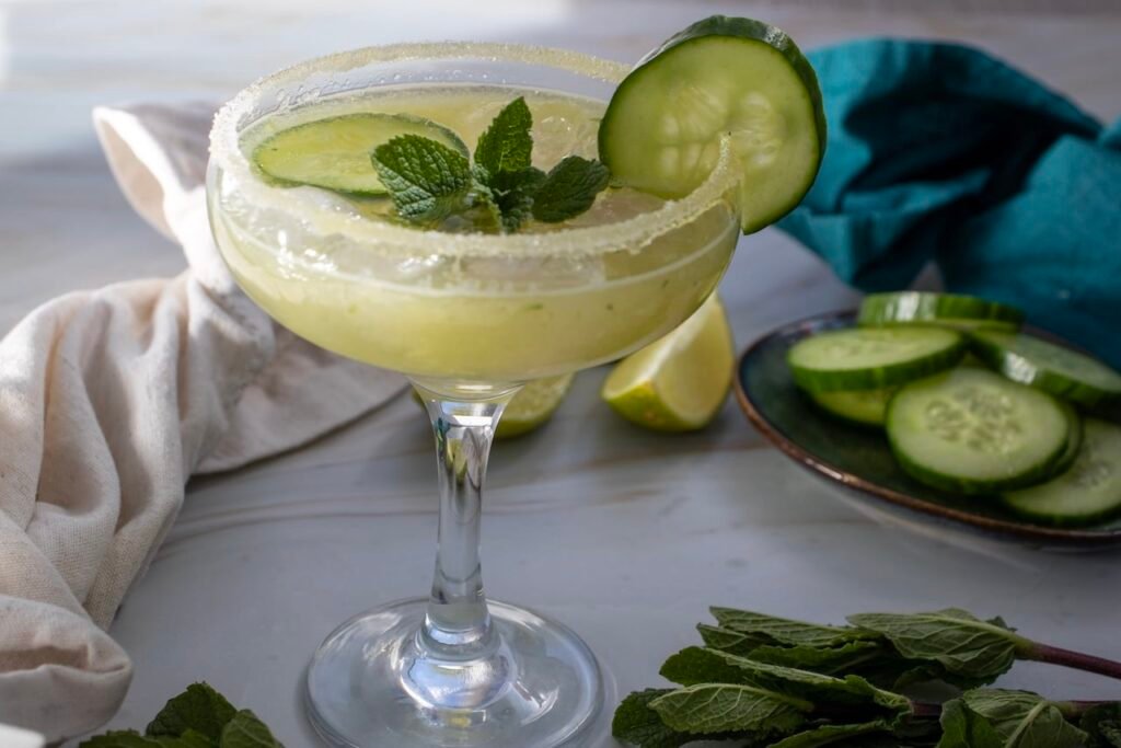 Cucumber margarita mocktail in a glass with a salted rim, garnished with cucumber slices and fresh mint, with lime wedges and cucumber slices in the background.