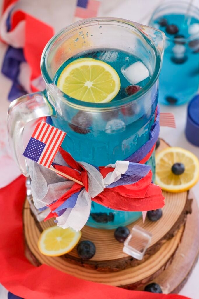Pitcher of blue 4th of July mocktail with lemon slice, blueberries, and ice, decorated with a red white and blue ribbon and small American flag.