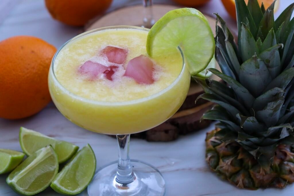 Pineapple margarita mocktail served over ice in a glass with lime garnish, surrounded by fresh pineapple, oranges, and lime wedges.