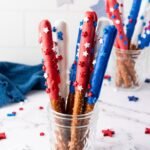 Firecracker pretzel rods dipped in red, white, and blue candy coating with star sprinkles, arranged upright in a glass jar on a light surface.