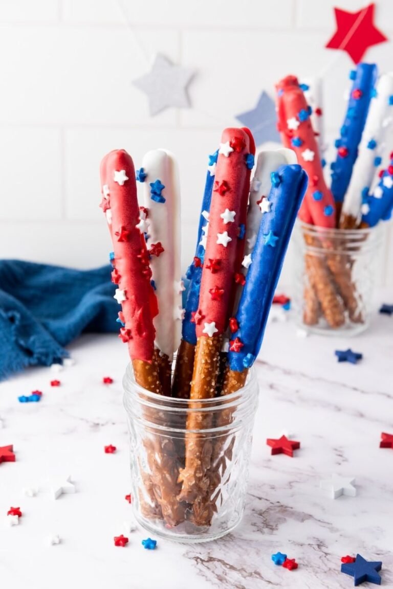 Firecracker pretzel rods dipped in red, white, and blue candy coating with star sprinkles, arranged upright in a glass jar on a light surface.