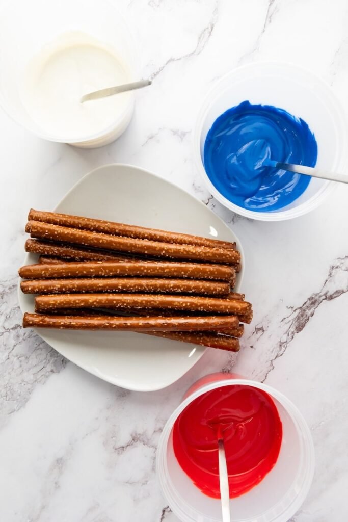 Pretzel rods on a plate next to bowls of melted red, white, and blue candy melts with spoons, showing the dipping setup for firecracker pretzel rods.