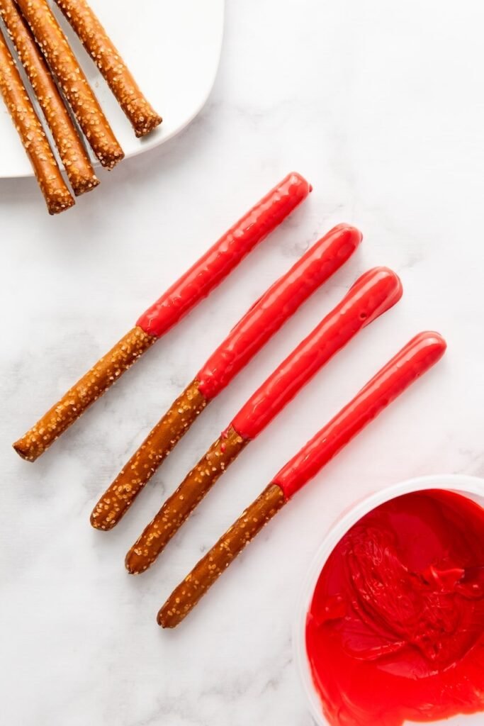 Close-up of a pretzel rod being dipped into melted red candy coating, showing the coating process for firecracker pretzel rods.