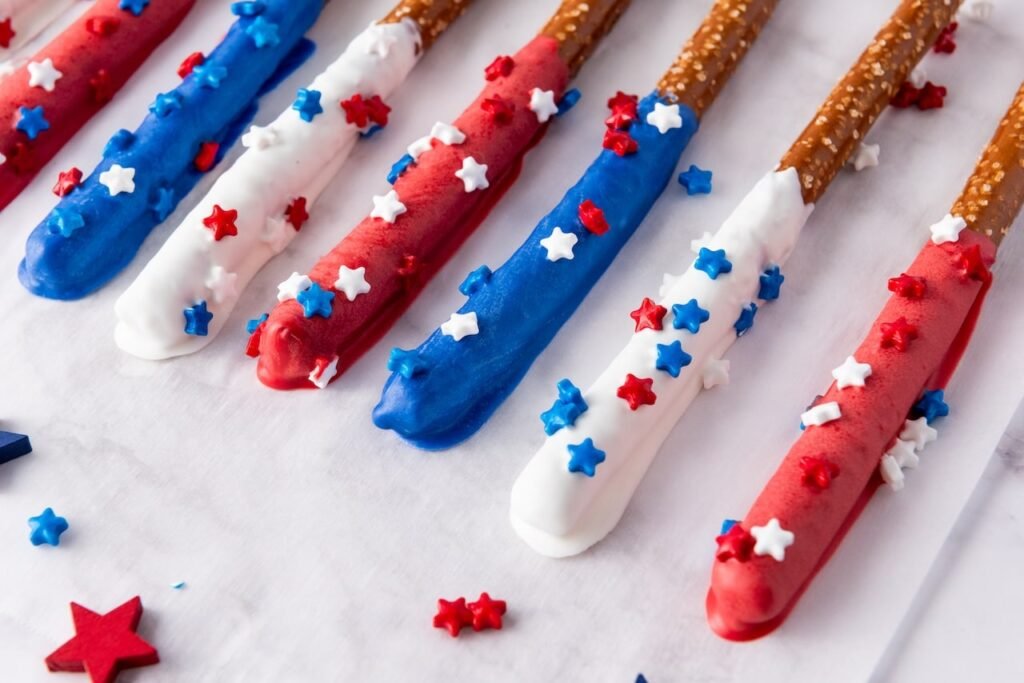 Close-up of firecracker pretzel rods dipped in red, white, and blue candy coating and topped with star sprinkles on a white surface.