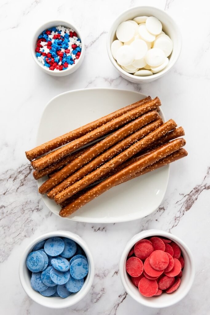Ingredients for firecracker pretzel rods including pretzel rods, red and blue candy melts, and patriotic sprinkles arranged on a countertop.