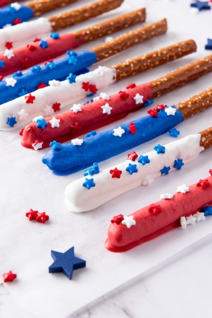 Candy-coated firecracker pretzel rods in red, white, and blue lined on a white surface and decorated with star sprinkles for a patriotic dessert.