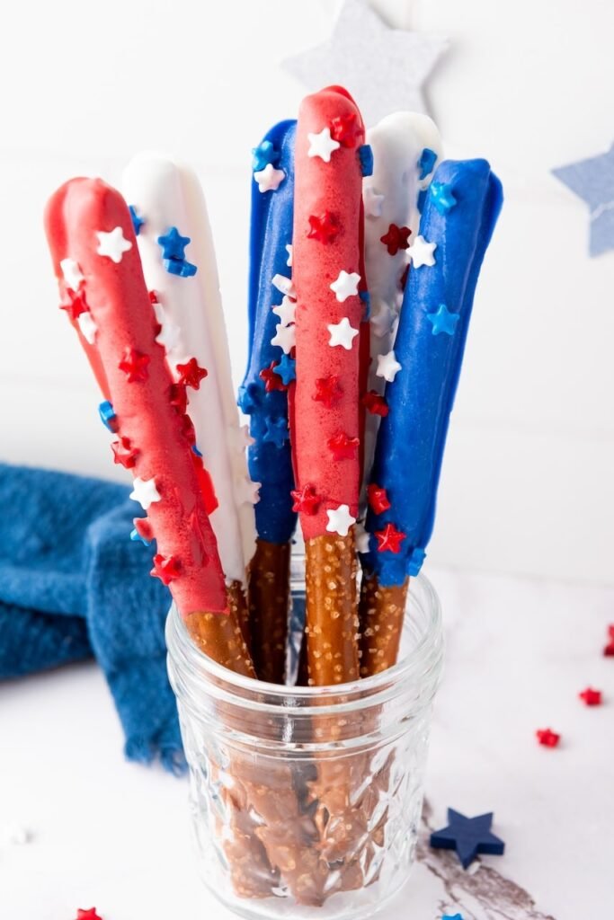 A glass jar filled with chocolate-coated firecracker pretzel rods decorated with red, white, and blue sprinkles, styled for a patriotic 4th of July dessert display.