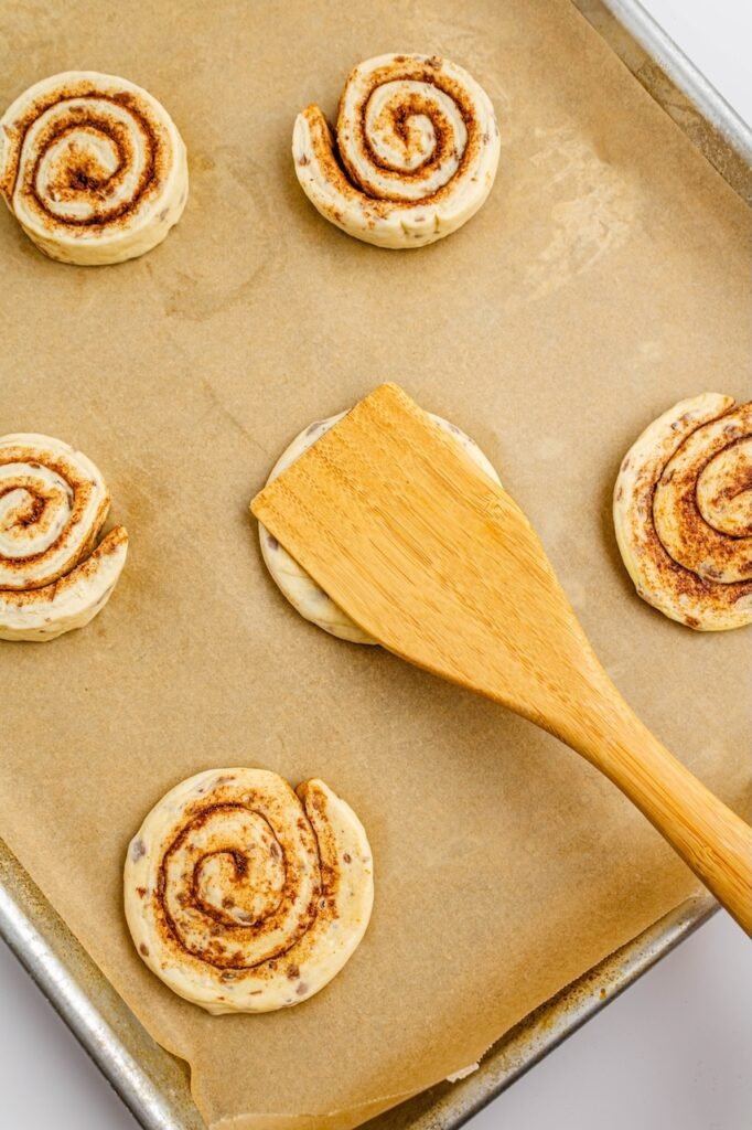 Overhead view of cinnamon rolls on parchment paper with a spatula pressing one roll flat to prepare it for cutting into a star shape.