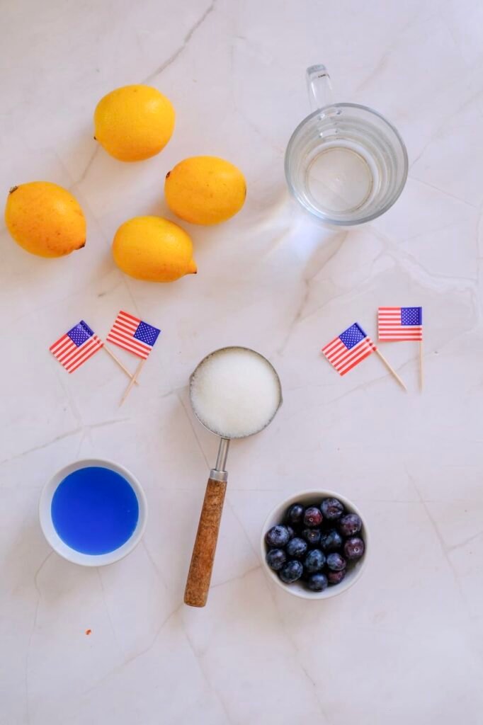 Fresh lemons, blueberries, sugar, blue curaçao syrup, and sparkling water arranged on a light countertop for a homemade recipe.