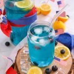 Blue 4th of July mocktail in a glass and pitcher with ice, lemon slices, and blueberries, styled with small American flags on a wooden board.