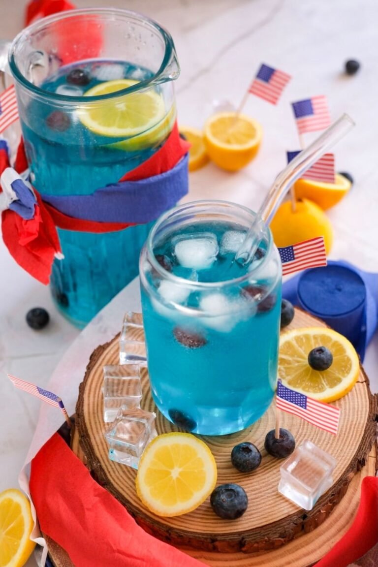 Blue 4th of July mocktail in a glass and pitcher with ice, lemon slices, and blueberries, styled with small American flags on a wooden board.