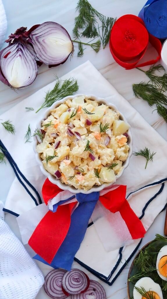 Top view of creamy potato salad in a white bowl garnished with dill, surrounded by red onion slices and red, white, and blue decorations.