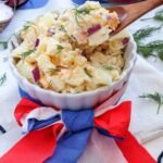 A bowl of creamy potato salad with chopped potatoes, eggs, red onion, and fresh dill, served in a white dish with red, white, and blue decorations for a Fourth of July meal.