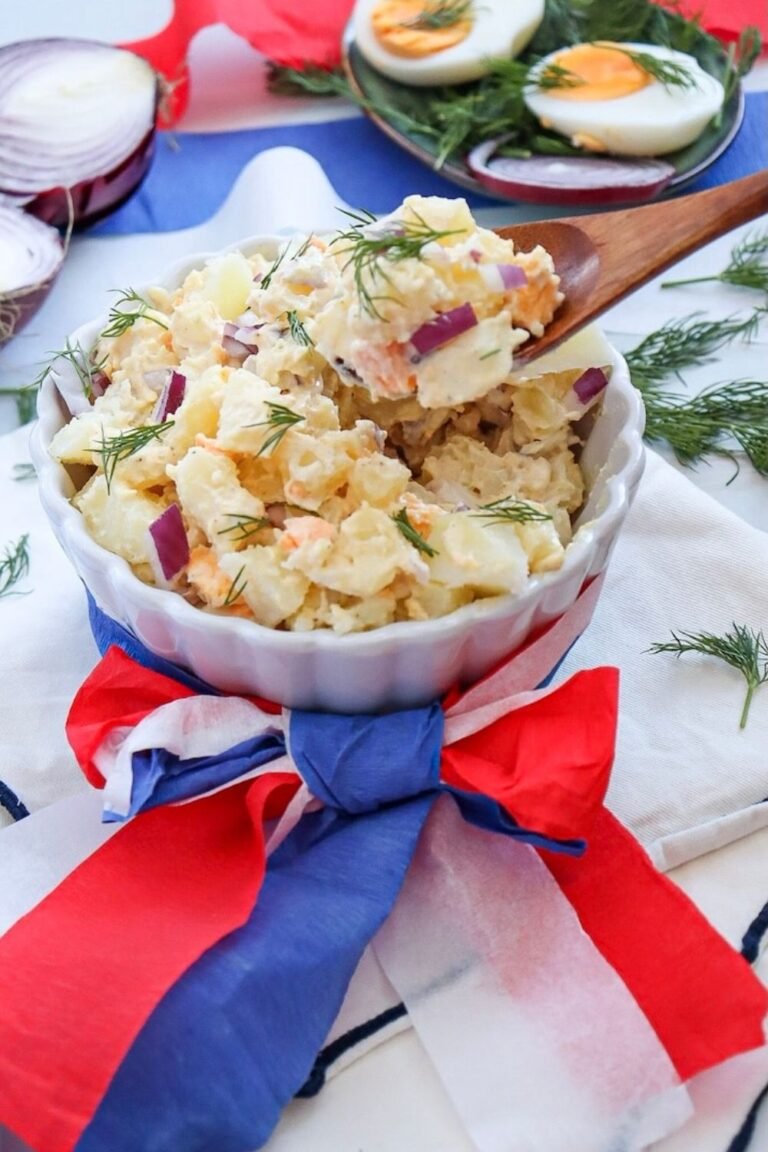 A bowl of creamy potato salad with chopped potatoes, eggs, red onion, and fresh dill, served in a white dish with red, white, and blue decorations for a Fourth of July meal.