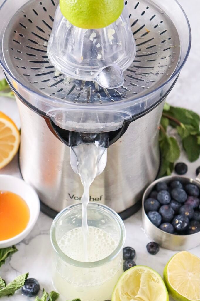 Fresh lime being juiced into a glass using a citrus juicer, with blueberries, mint, and honey nearby for a blueberry mojito mocktail.