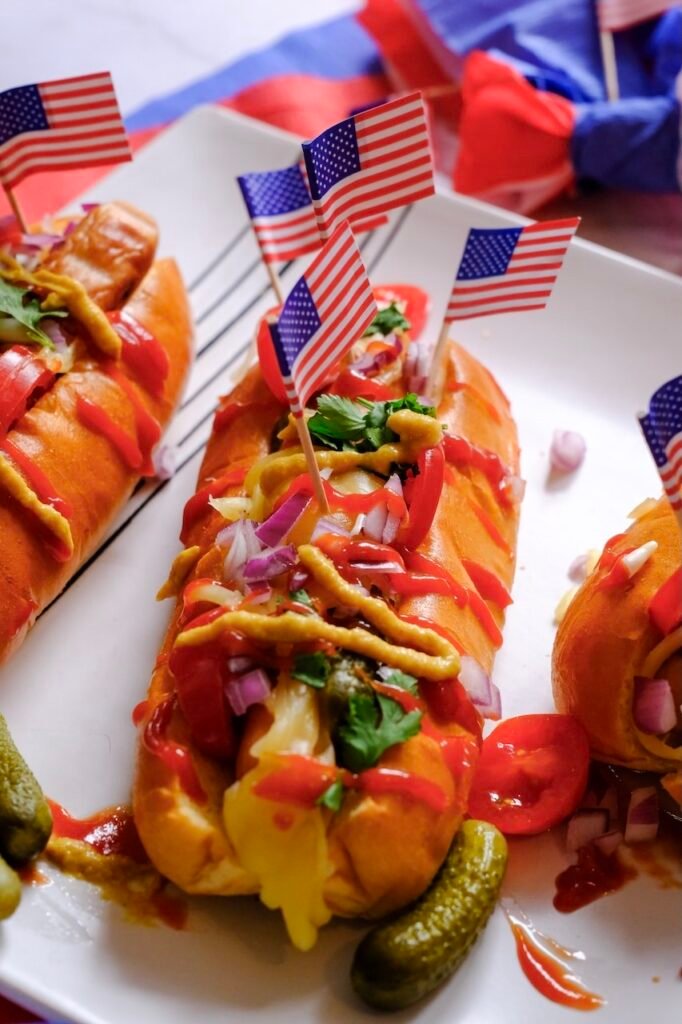 Close-up of grilled hot dogs in buns topped with melted cheese, chopped onions, pickles, and condiments, decorated with small American flags for a summer cookout.