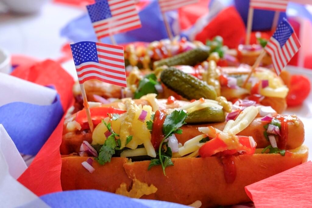 Close-up of grilled hot dogs in buns topped with pickles, chopped onions, cheese, and condiments, decorated with small American flags for a summer cookout.