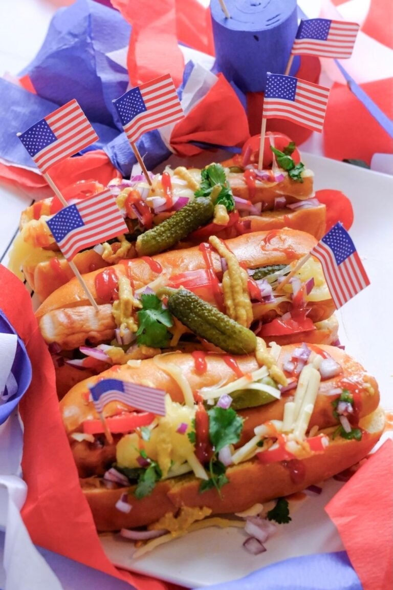 Grilled hot dogs in buns topped with pickles, onions, cheese, and condiments, decorated with small American flags for a July 4th cookout.
