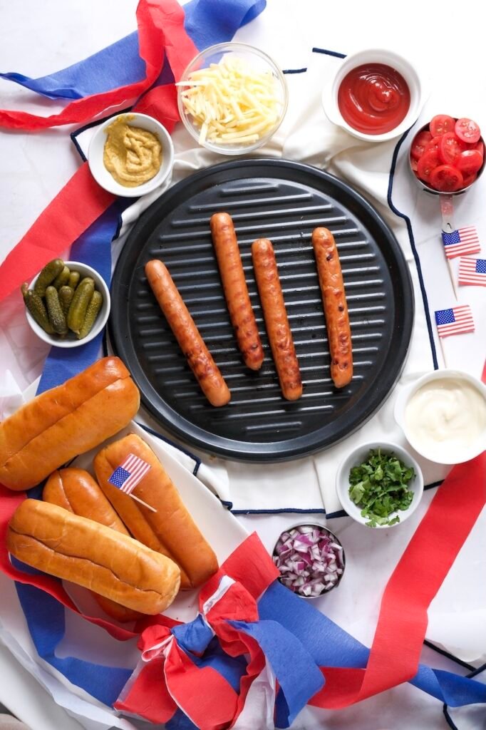 dogs cooking on a ridged grill pan with visible grill marks, served with pickles on the side during a summer cookout setup.