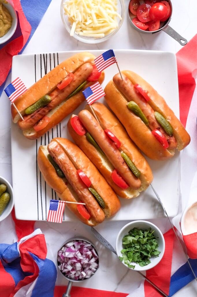 dogs with pickles and toppings arranged on a plate for a summer cookout, with small American flags for decoration.