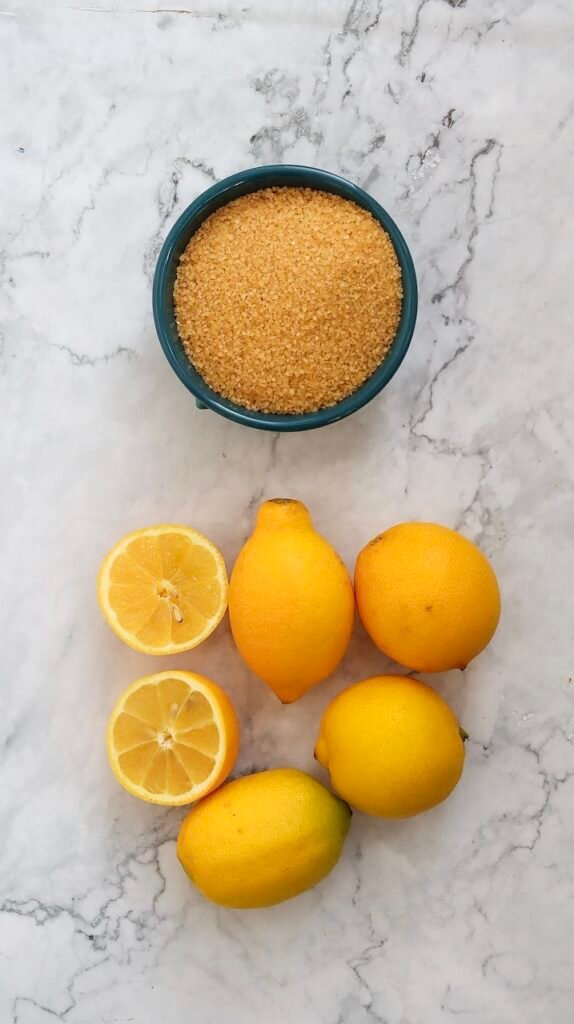 Overhead view of fresh lemons, halved lemons, and a bowl of granulated sugar on a marble surface, showing the ingredients needed to make homemade lemonade.