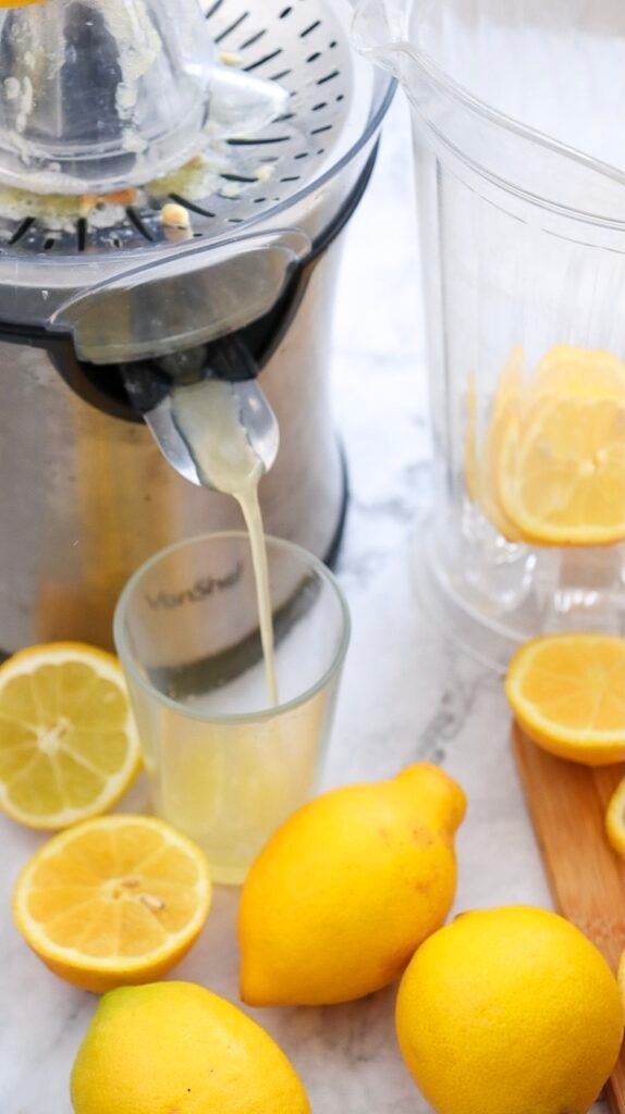 Fresh lemon juice being pressed with a juicer into a glass, surrounded by whole and halved lemons, showing how to prepare lemon juice.