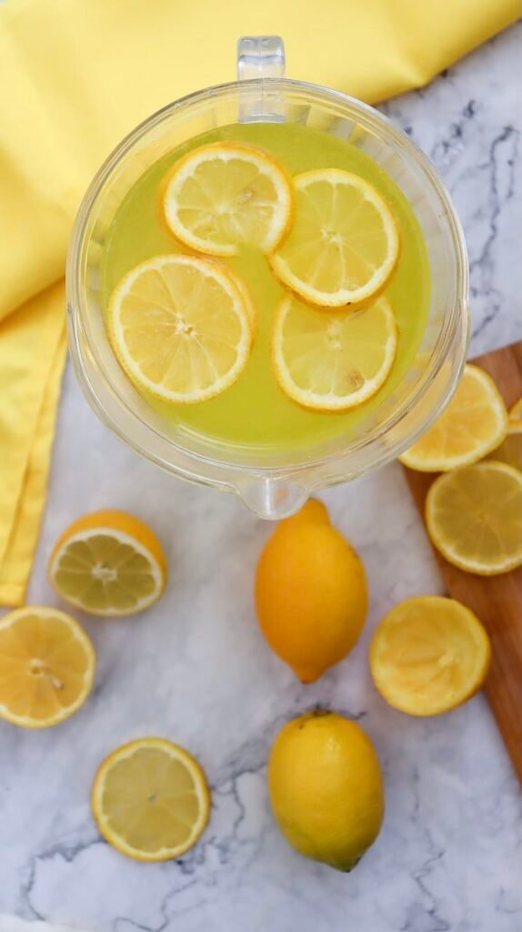 Overhead view of a pitcher filled with homemade lemonade and floating lemon slices, surrounded by fresh whole and sliced lemons, showing the finished lemonade ready to serve.