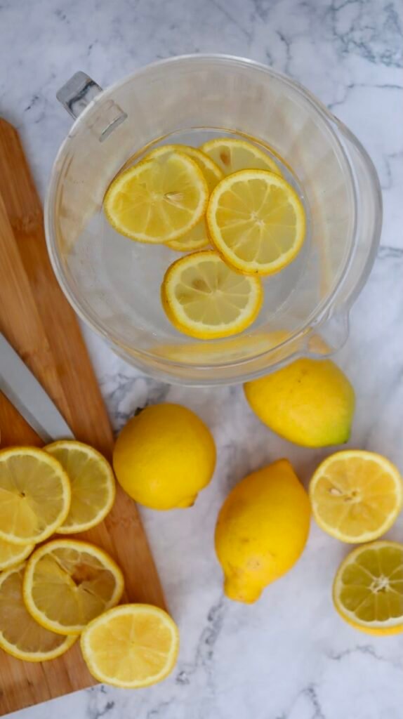 Overhead view of a clear pitcher with lemon slices in water, surrounded by fresh whole and sliced lemons on a marble surface, showing preparation.