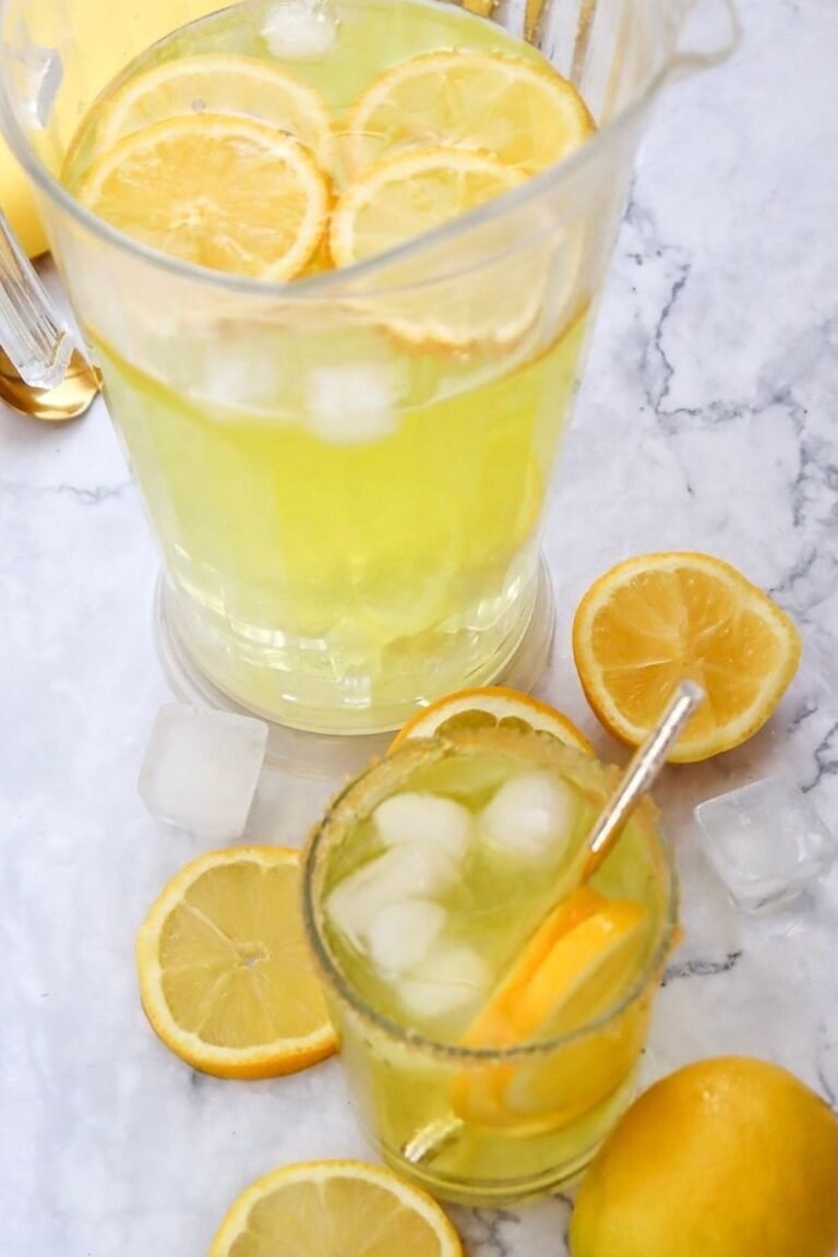 A glass pitcher and a small glass filled with homemade lemonade over ice, garnished with lemon slices, showing the finished drink ready to serve on a marble surface.