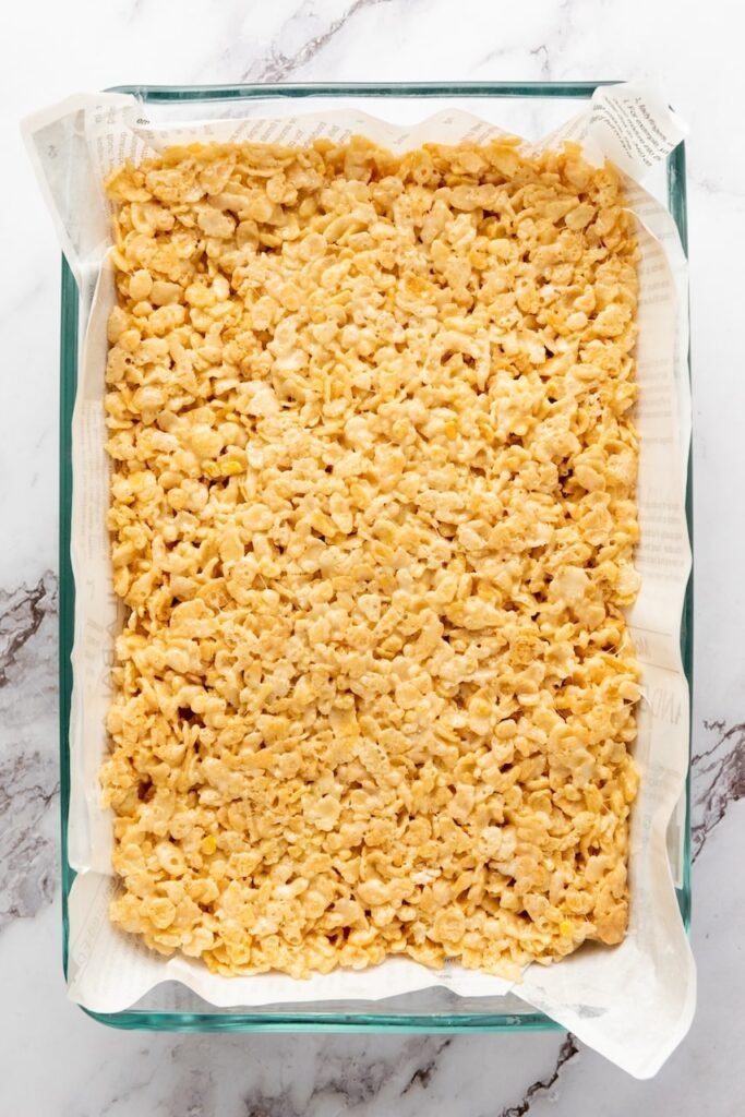 Overhead view of cereal snack mixture pressed evenly into a parchment-lined baking dish, ready to set before cutting into squares.