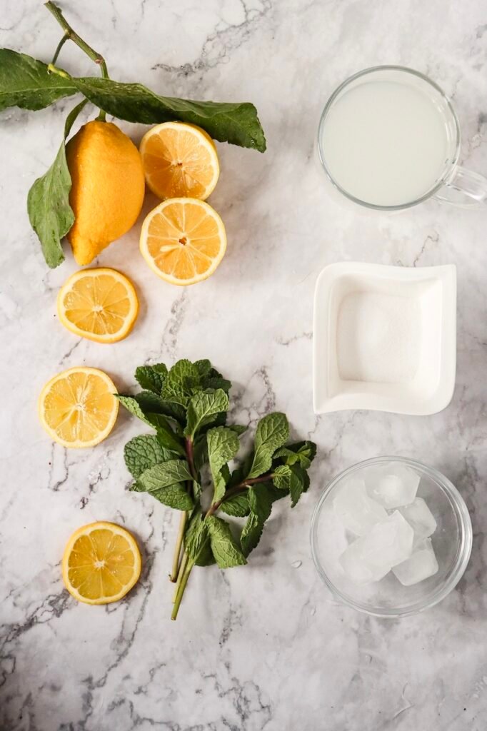 Overhead view of fresh lemons, sliced lemon rounds, mint sprigs, ice cubes, sugar, and a glass of lemon juice arranged on a marble surface for a lemon mojito mocktail recipe.