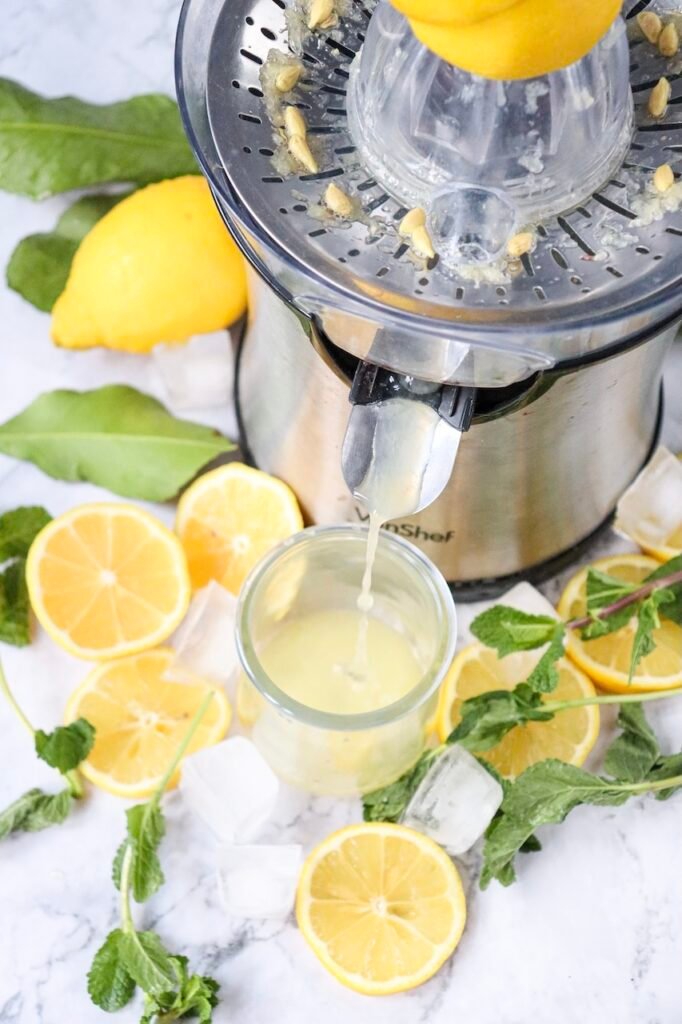 Close-up of fresh lemons being juiced with an electric juicer into a glass, surrounded by mint leaves, lemon slices, and ice for a virgin citrus drink recipe.