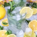 Two glasses of lemon mojito mocktail with ice, fresh mint, and lemon slices, surrounded by sliced lemons and ice on a marble surface.