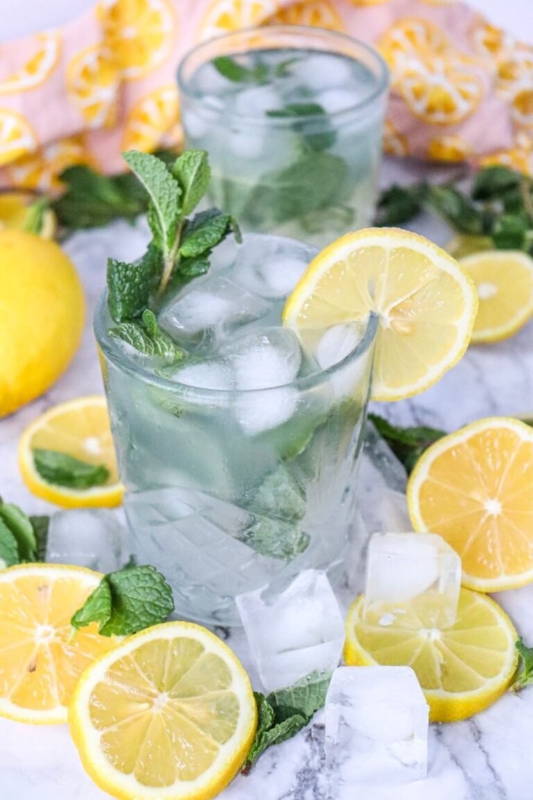 Two glasses of lemon mojito mocktail with ice, fresh mint, and lemon slices, surrounded by sliced lemons and ice on a marble surface.