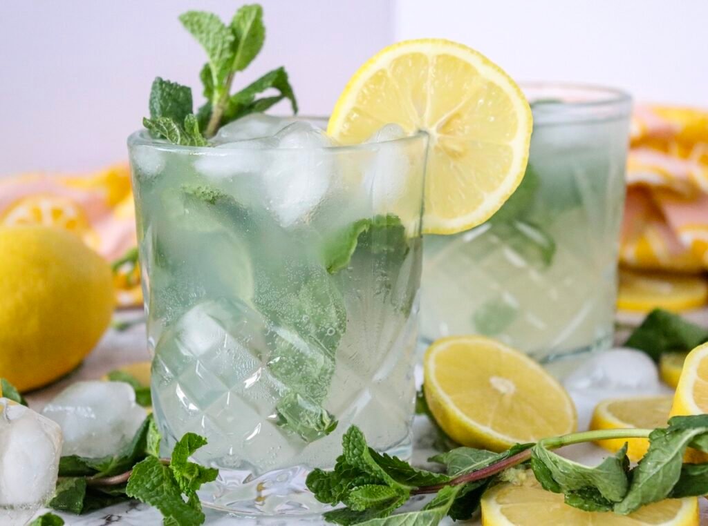 Close-up of a lemon mojito mocktail in a glass with ice, fresh mint leaves, and a lemon slice garnish, with a second glass in the background.