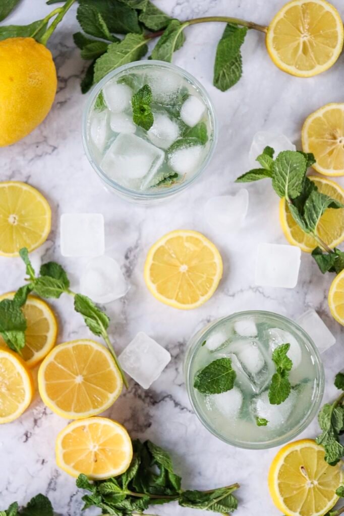 Overhead view of two glasses of lemon mojito mocktail with ice and mint, surrounded by sliced lemons, fresh mint sprigs, and ice cubes on a marble surface.