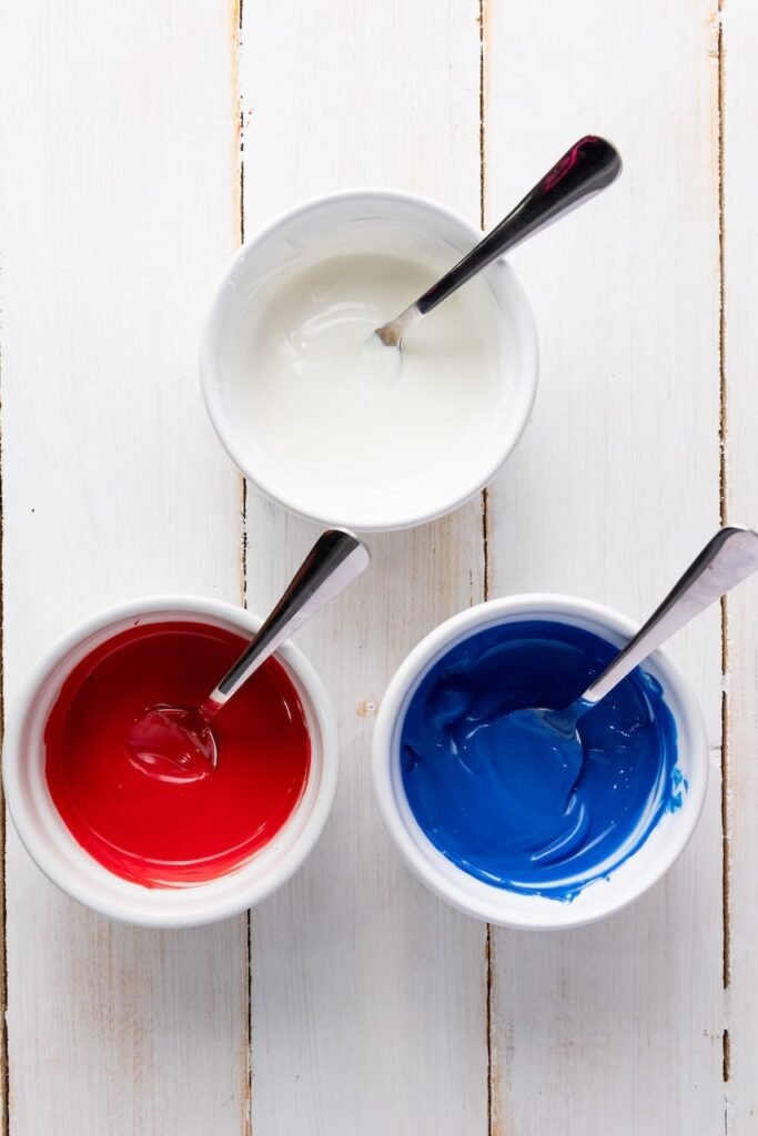 Three small bowls filled with melted candy melts in red, white, and blue, each with a spoon, sitting on a white wooden surface, ready to be used for drizzling over popcorn.