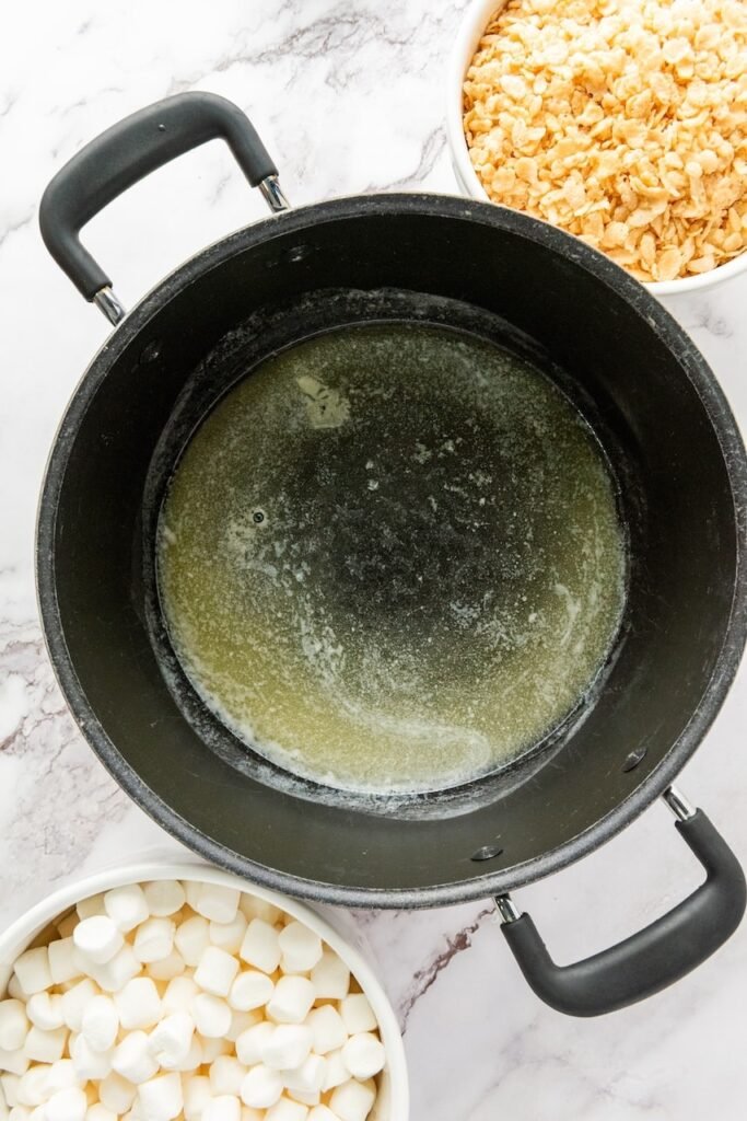 Overhead view of melted butter in a large pot on the stove, with bowls of rice krispies cereal and mini marshmallows nearby for making rice krispie treats.