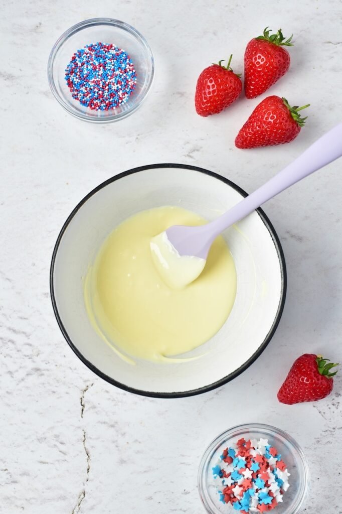 Melted white chocolate in a bowl being stirred with a spatula, with strawberries and red, white, and blue sprinkles nearby for a holiday dessert.