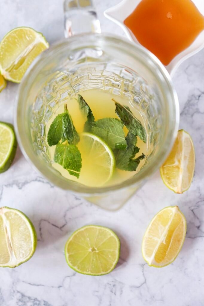 Overhead view of a glass pitcher with lime slices and fresh mint leaves in a partially mixed base on a marble surface.