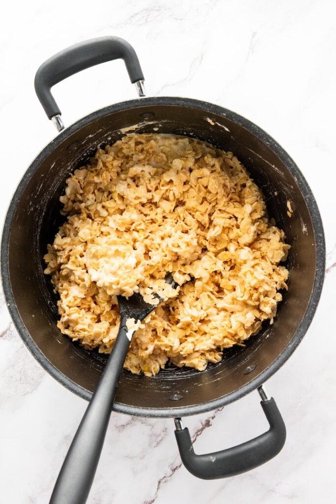 Overhead view of cereal snack being mixed into melted marshmallow mixture in a pot with a spatula.