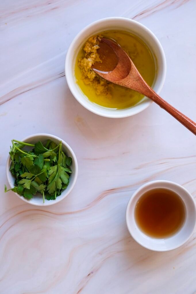 Close-up of olive oil and Dijon mustard being mixed in a small bowl with a wooden spoon, with parsley and red wine vinegar nearby for homemade dressing.