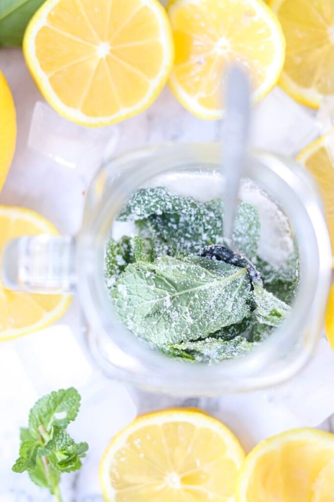 Top view of fresh mint leaves and sugar being muddled in a glass with a spoon, surrounded by sliced lemons, for a virgin citrus drink preparation step.