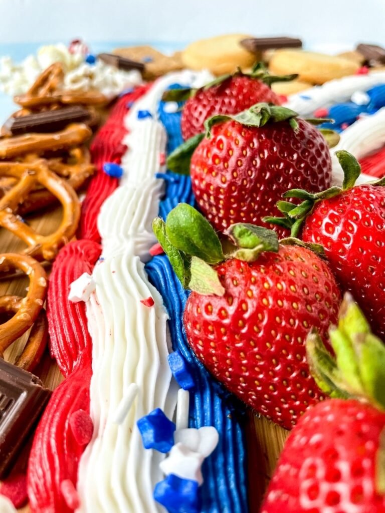 Close-up view of fresh strawberries lined along red, white, and blue buttercream frosting with pretzels and chocolate on a wooden dessert board.