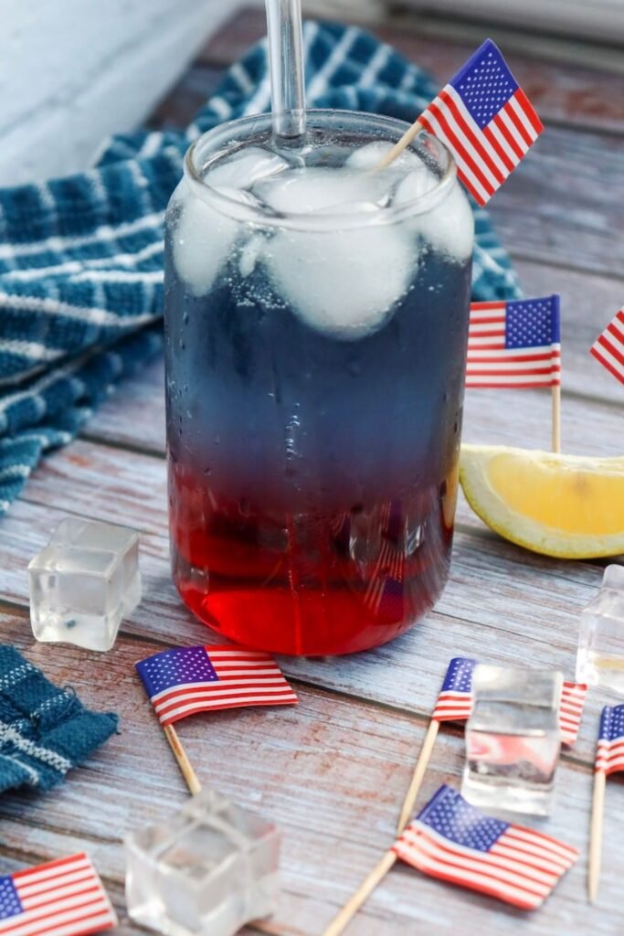 Glass of layered patriotic punch showing red, white, and blue layers with ice, a straw, lemon slice, and small American flags on a wooden surface.