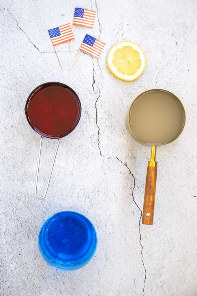Top view of ingredients for patriotic punch including cranberry juice, lemonade, blue sports drink, a lemon slice, and small American flags arranged on a light surface.