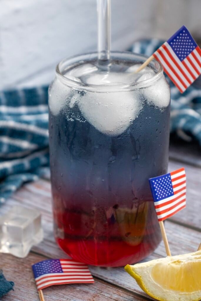 Close-up of a glass of layered patriotic punch with red, white, and blue layers, ice cubes, a straw, lemon wedge, and small American flags on a wooden table.