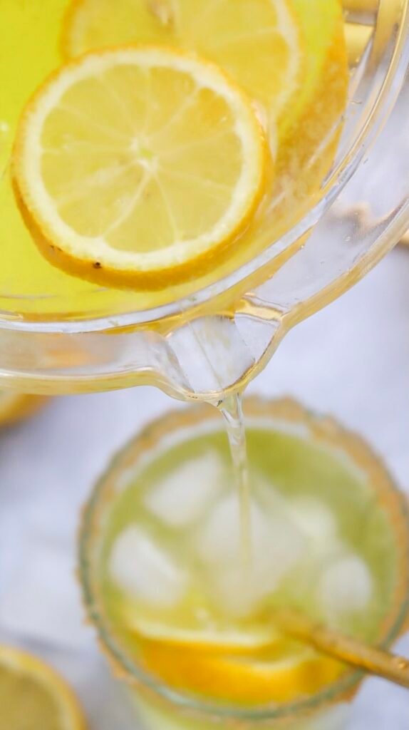 Close-up of homemade lemonade being poured from a pitcher into a glass filled with ice and lemon slices, showing the final step of serving the drink; overlay text reads “Perfect Homemade Lemonade.”