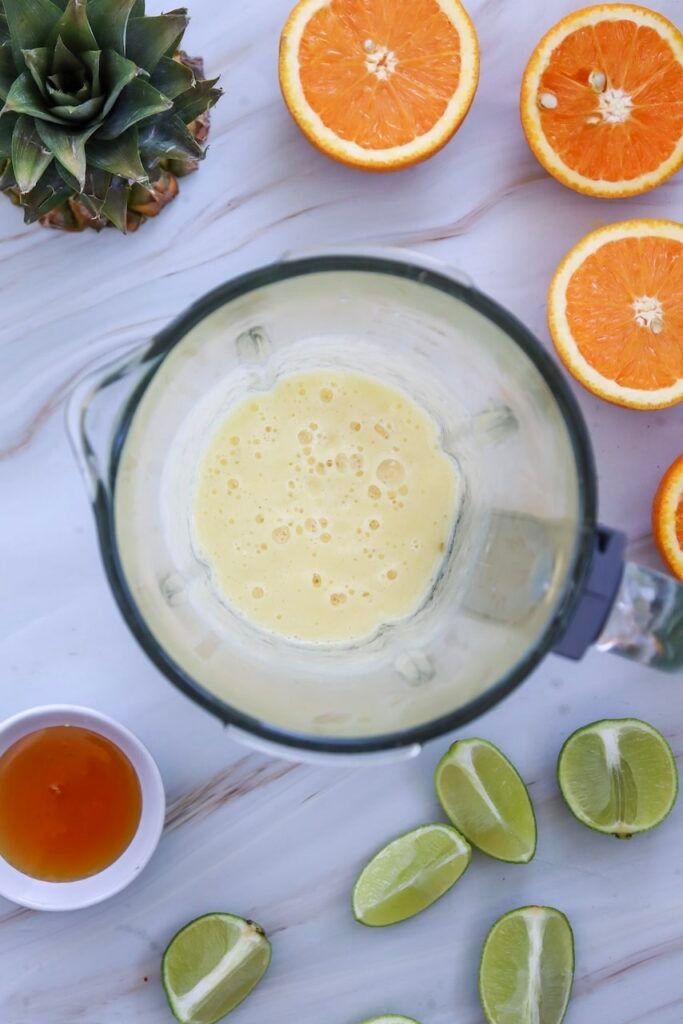 Blended pineapple mixture in a blender with fresh limes, orange slices, and honey nearby, showing the smoothie-like base for a pineapple margarita mocktail.