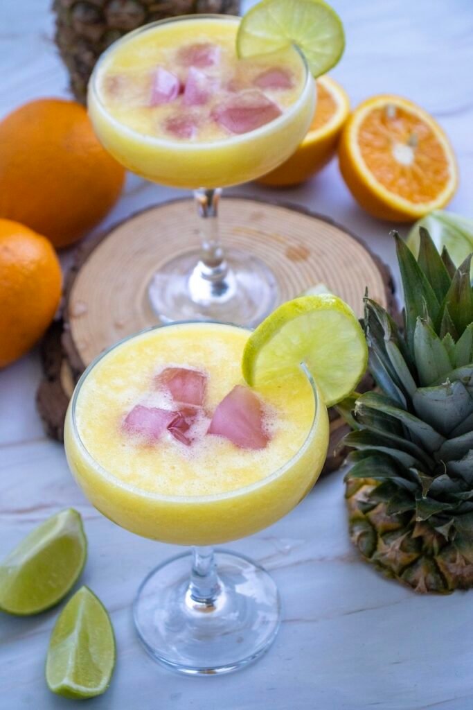 Two pineapple margarita mocktails served over ice in glasses with lime garnish, surrounded by fresh pineapple, oranges, and lime wedges.